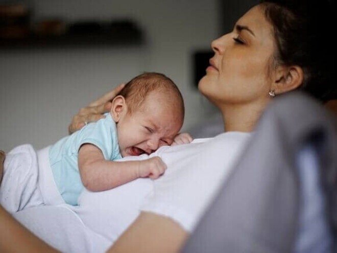 Uma mãe com aspecto de cansaço, deitada com um bebê chorando em seu colo.