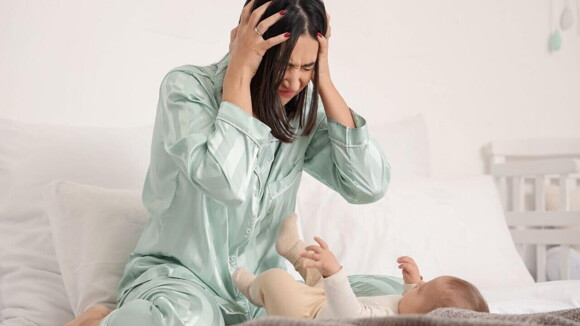 Uma mãe com as mãos na cabeça e aspecto de desespero, sentada na cama, em frente de um bebê deitado.